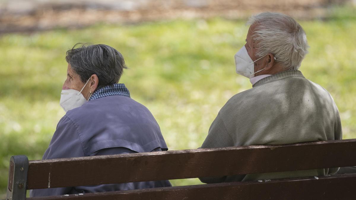 Un hombre y una mujer mayores, sentados en un banco y con mascarilla.