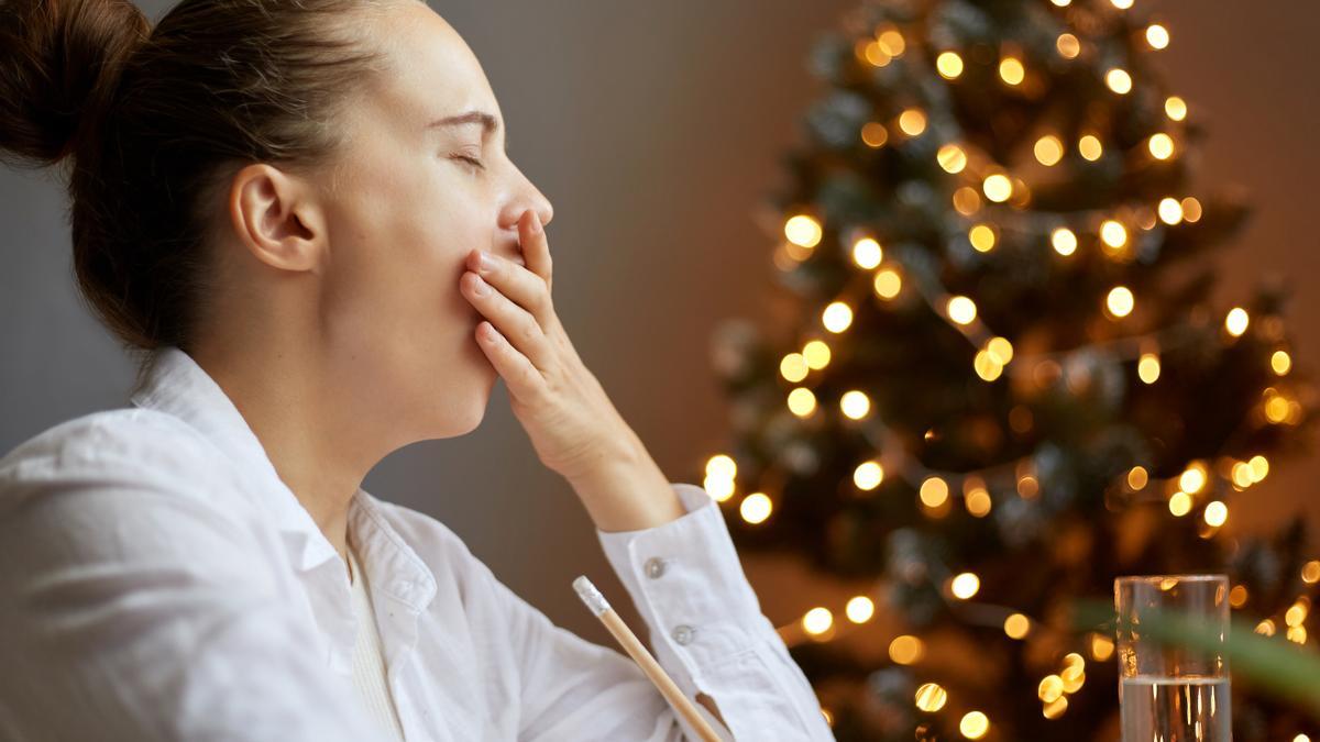 Portrait of tired sleepy woman wearing white shirt posing in decorated room near Christmas tree, has not enough sleep, yawning, covering mouth with her palm.