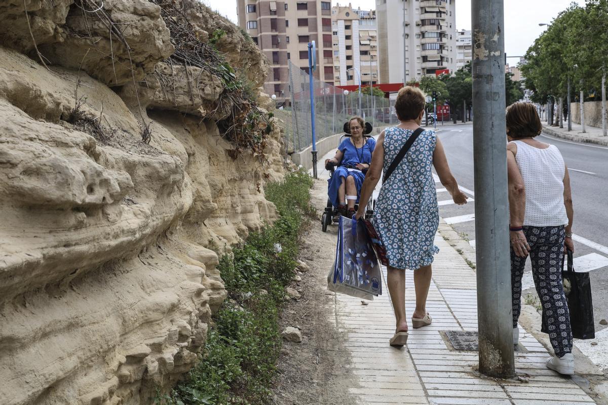 Varias vecinas de la zona pasean por una acera ocupada por farolas, tierra y maleza.