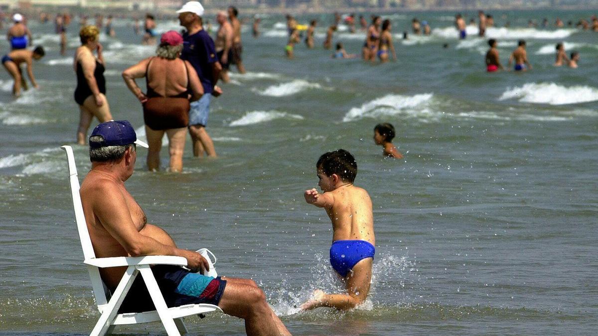 Un niño juega en la orilla de la playa Las Arenas de Valencia mientras un hombre se refresca las piernas sentado a su lado.