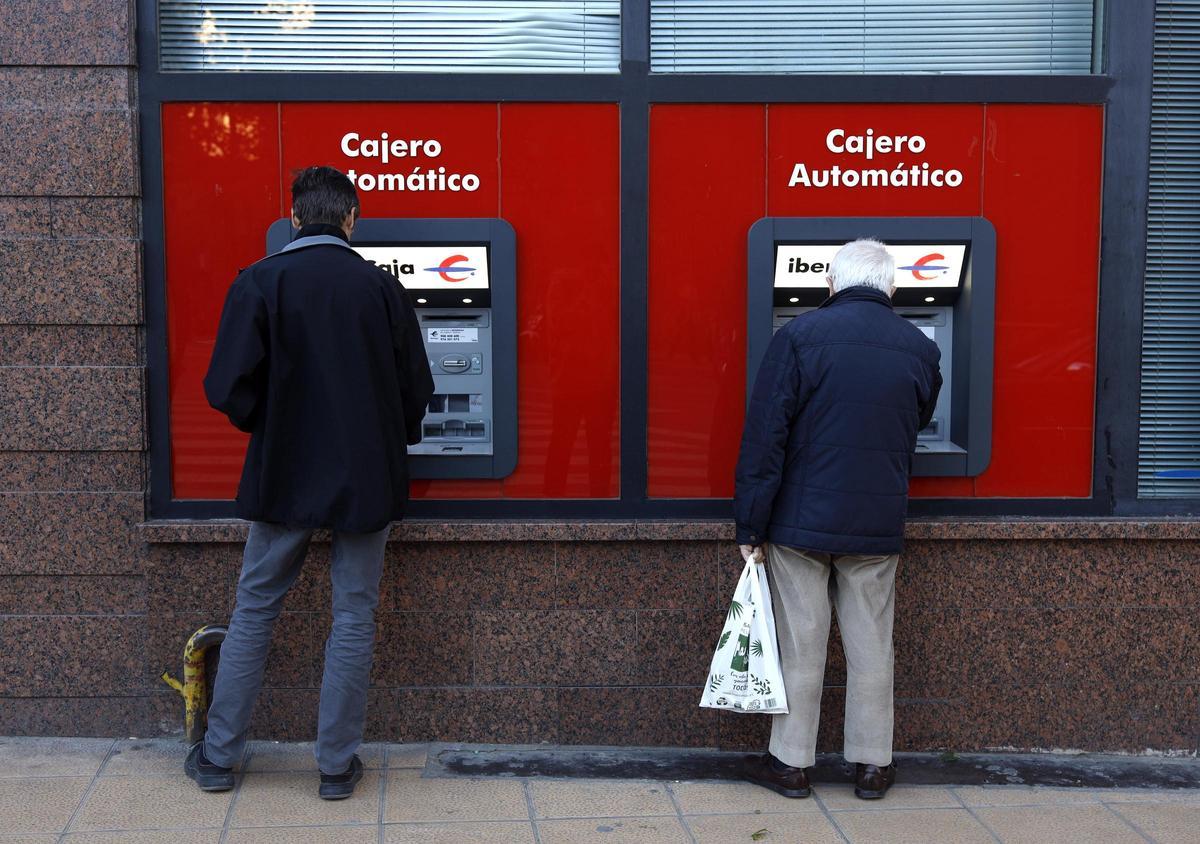 Dos personas utilizan sendos cajeros automáticos en una sucursal bancaria.