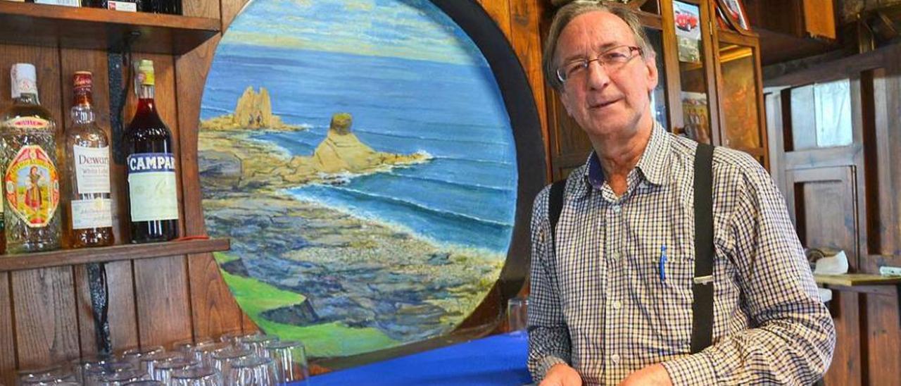 Macario Fernández Fernández, tras la barra de su establecimiento, junto a una de sus pinturas, en la que se ve la playa de Portizuelo.