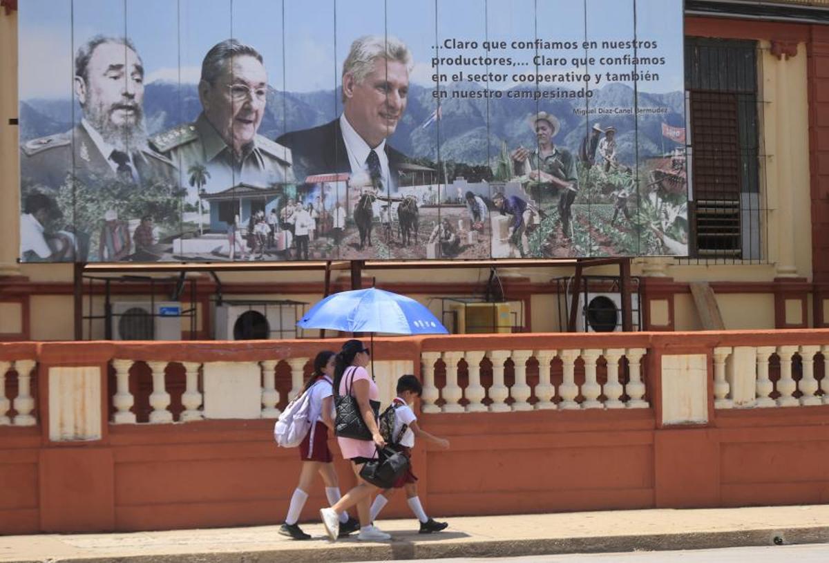Una madre y sus dos hijos caminando por La Habana delante de un mural con los líderes de la revolución cubana.