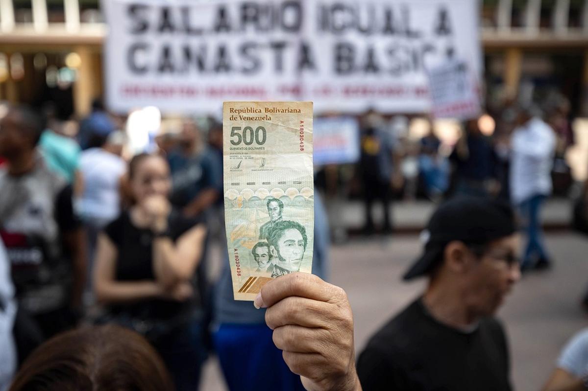 Un manifestante sostiene un billete de quinientos bolívares, al cambio aproximadamente un euro, durante una protesta frente al Ministerio de Trabajo en Caracas para exigir un aumento de salarios y pensiones en Venezuela. Detrás, una pancarta pide "salario igual a canasta básica".