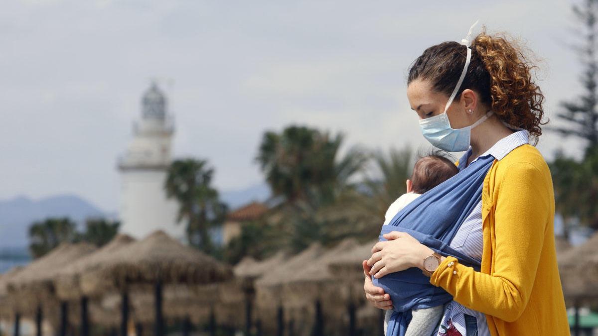 Una mujer protegida con una mascarilla da un paseo con su bebé en la playa de La Malagueta