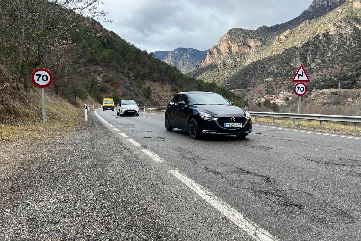 Diversos vehicles circulant per un tram de la carretera N-260 ple de clots, on es veu el poble del Pont de Bar (Alt Urgell) al fons