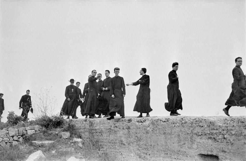 Descontextualitzar ha sigut sempre un exercici d'exploració pels fotògrafs. L'escena parteix d'un element totalment reconeixible, un grup de sacerdots dels germans Maristes amb sotana, que el fotògraf fotografia en un entorn que en principi no els correspon, un mur de les ruïnes d'Empúries. Veure els protagonistes en un rol que no els és habitual, el lleure, proporciona una imatge molt suggeridora per l'espectador que veu un component d'humanització en aquestes figures destinades al servei diví.