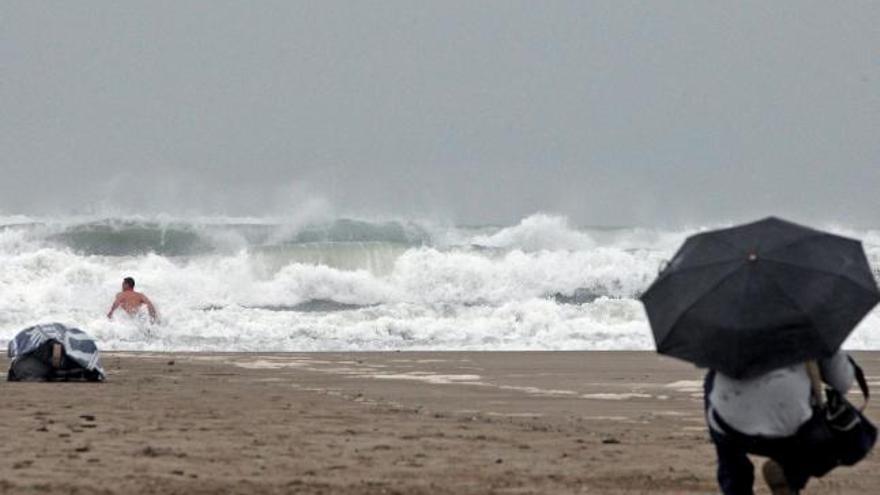 Un hombre protegido con un paraguas observa como otro se baña entre un fuerte oleaje este mediodía en la playa de Las Arenas de Valencia