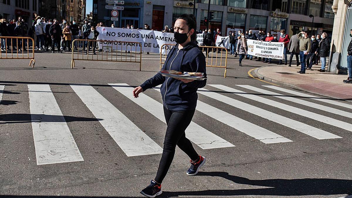 Una trabajadora de una terraza cruza la calle. Al fondo, los hosteleros protestan contra la Junta. | Emilio Fraile