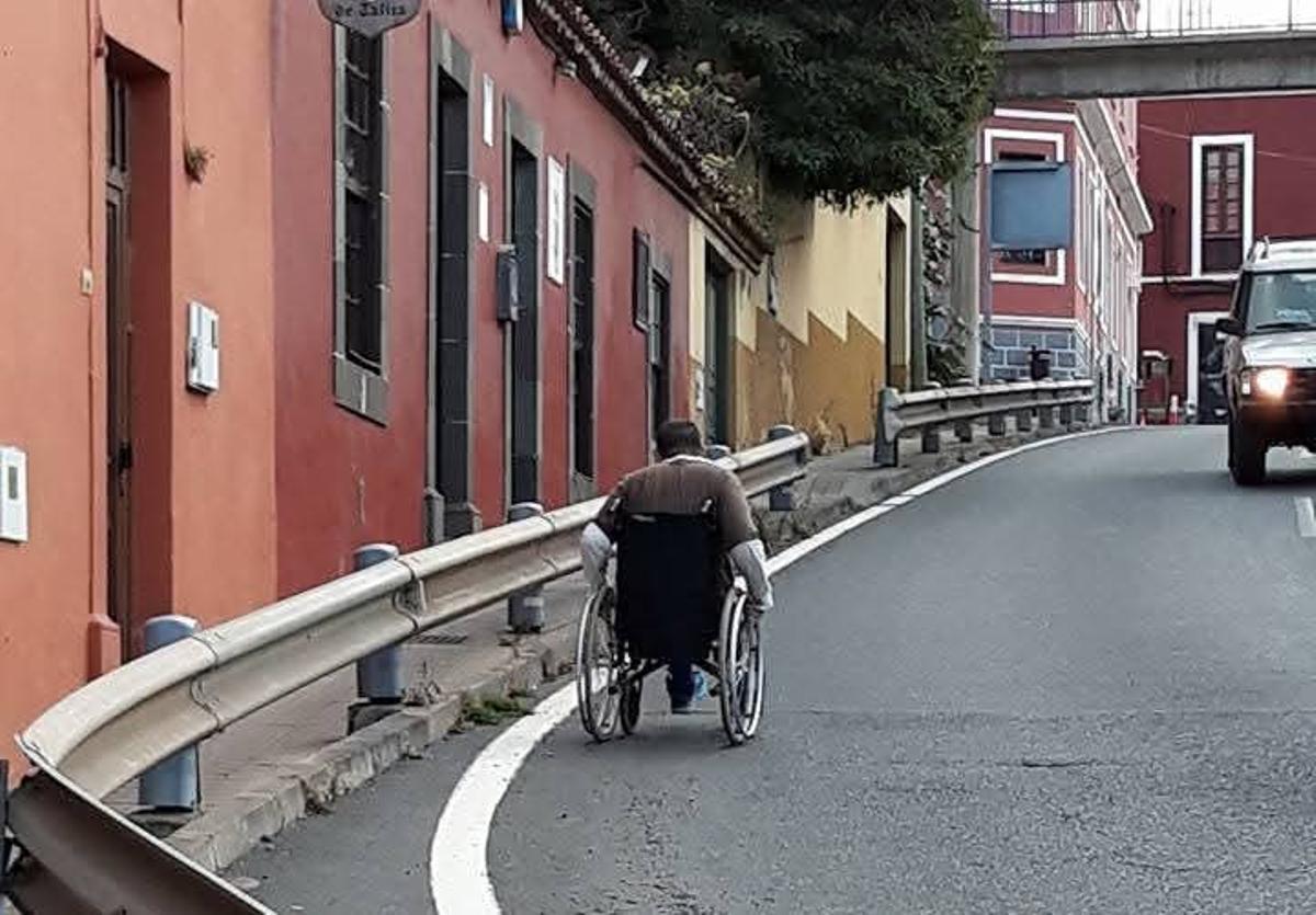 Un vecino con movilidad reducida va por la carretera debido al estrechamiento de la acera por la presencia de pivotes y vallas.