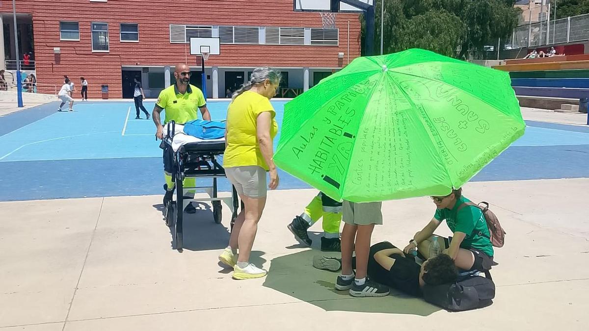 El alumno que se lesionó en el patio del IES  Cap de l'Horta bajo una sombrilla mientras le refrescan con agua por las altas temperaturas en el centro