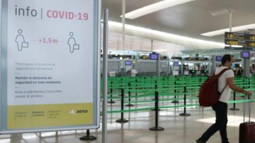 Imatge d&#039;arxiu d&#039;un cartell informatiu de la covid-19 i d&#039;un passatger arrossegant una maleta amb el control de seguretat de la T1 de l&#039;Aeroport del Prat