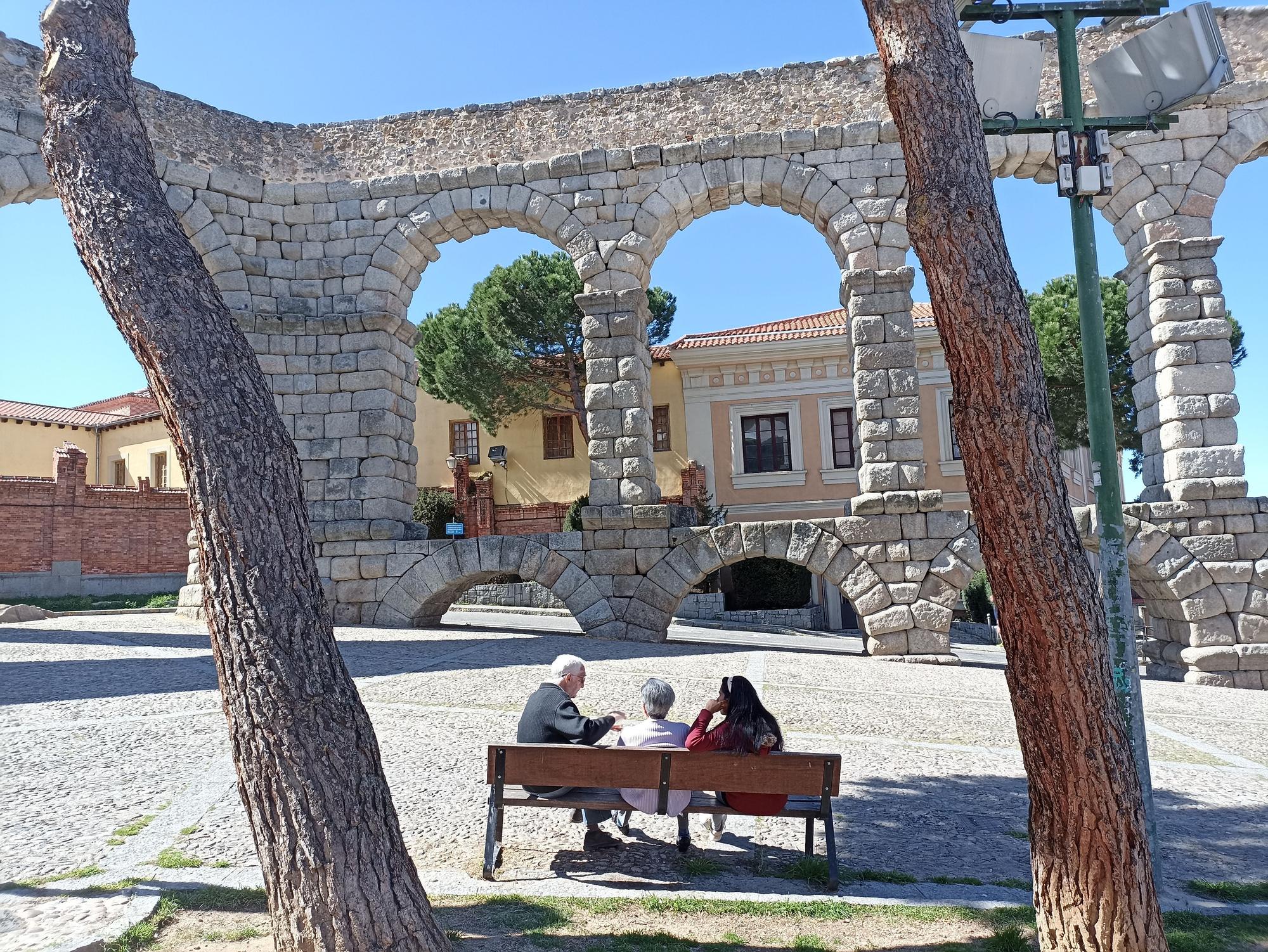 Varios segovianos charlan en un banco frente al punto del acueducto donde se suban más turistas.