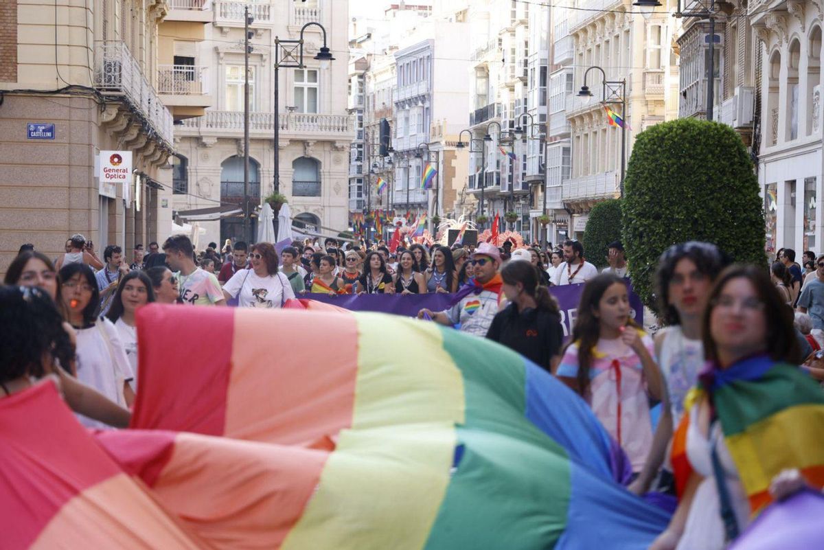 1. Miles de manifestantes pasan por la Puerta de Murcia con una bandera arcoirís. 2. Dos mujeres se besan subidas a una carroza. 3. Una mujer marcha con un niño portando una bandera del orgullo. 4. La carroza del partido socialista llena de gente desfila por las calles de Cartagena este sábado. | IVÁN URQUIZAR