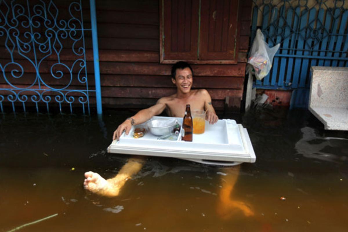 Un jove tailandès disfruta de la seva beguda davant de la porta de casa seva, que està inundada, en un suburbi de Bangkok (Tailàndia).