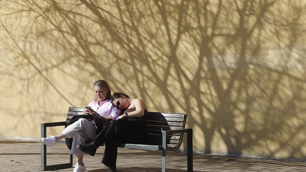 Dos jóvenes se sientan al sol en un banco de la avenida de la Ribera.