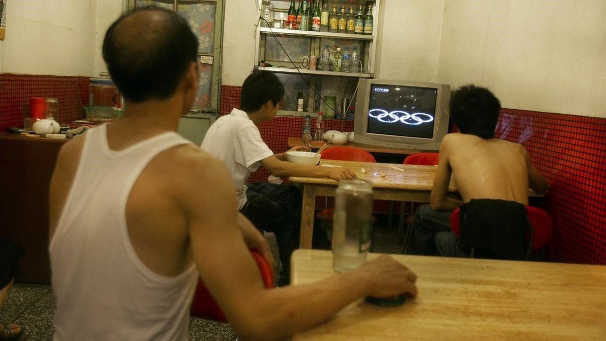 Tres chinos, alguno sin camiseta, siguen los juegos olímpicos a través de la televisión de un bar