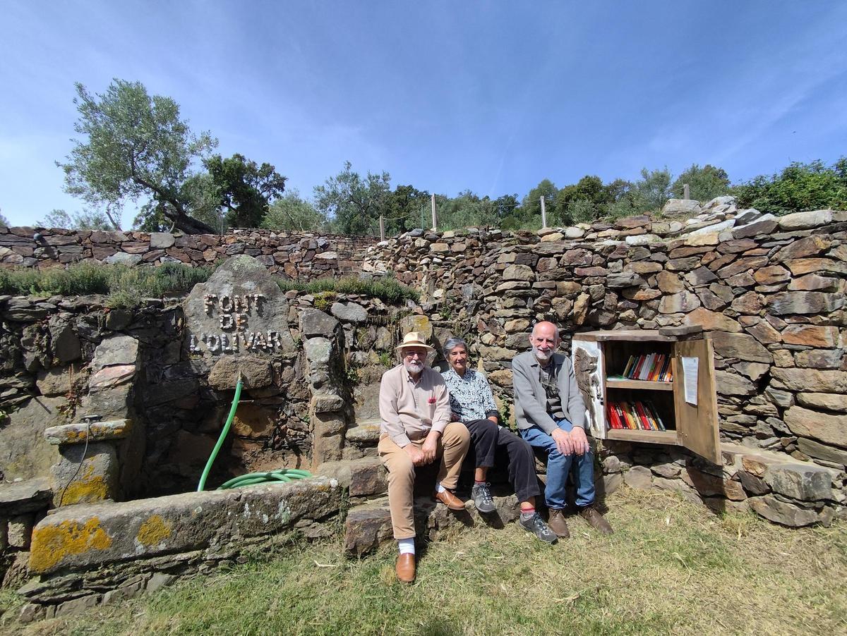 Tres dels impulsors de la biblioteca de muntanya al costat de la Font de l'Olivar i d'un dels mobles on els visitants poden trobar llibres.