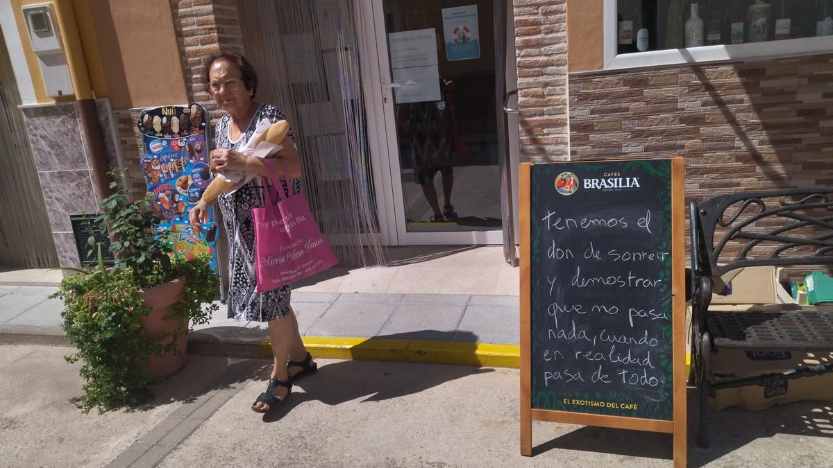 Una vecina sale con el pan de la Tendeta, en cuya puerta está la pizarra con el mensaje del &quot;don de sonreír&quot;