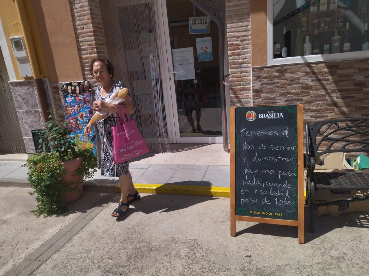Una vecina sale con el pan de la Tendeta, en cuya puerta está la pizarra con el mensaje del "don de sonreír"