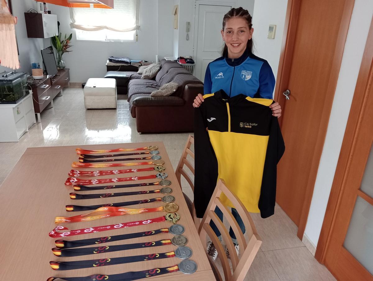 Adriana, vestida del CT Gandia, posa con una sudadera del CA Safor ante las medallas que ha ganado