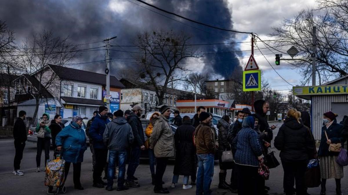 Ciudadanos ucranianos hacen fila frente a un supermercado mientras el humo se eleva sobre la ciudad de Vasylkiv, en las afueras de Kiev, este 27 de febrero de 2022, después de que los ataques rusos durante la noche golpearan un depósito de petróleo.