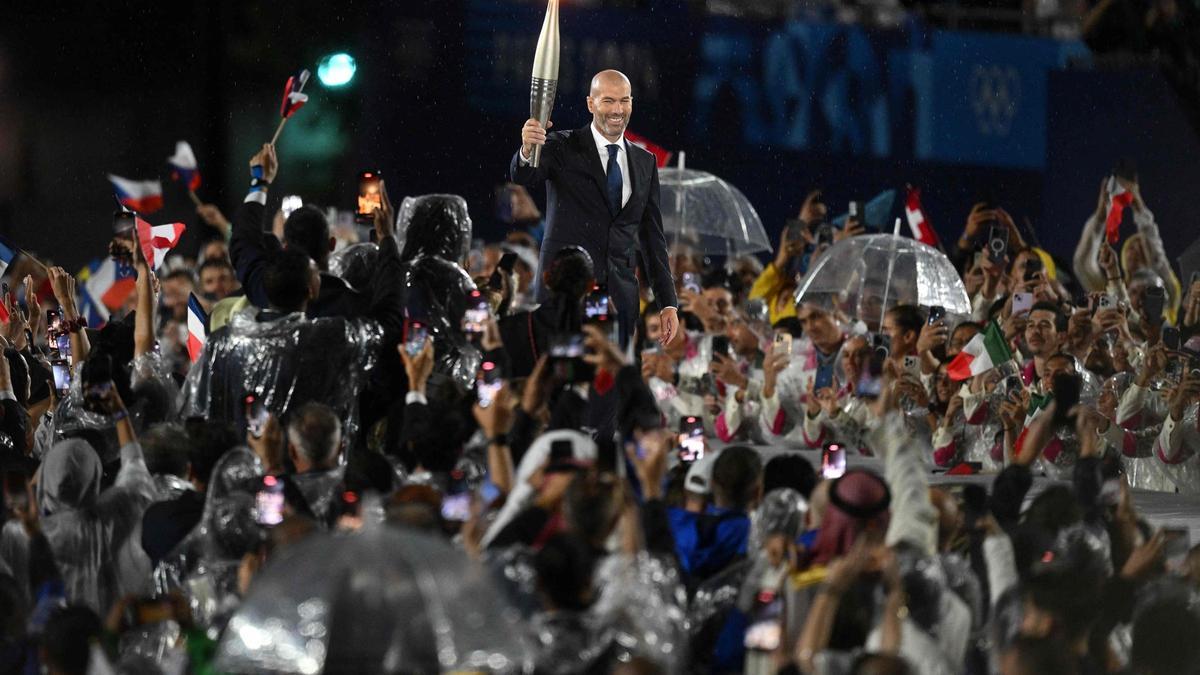 Zidane con la llama olímpica en el escenario arropado por deportistas con paraguas y chubasqueros.