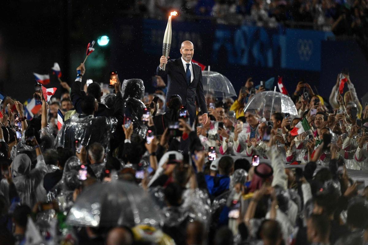 Zidane con la llama olímpica en el escenario arropado por deportistas con paraguas y chubasqueros.