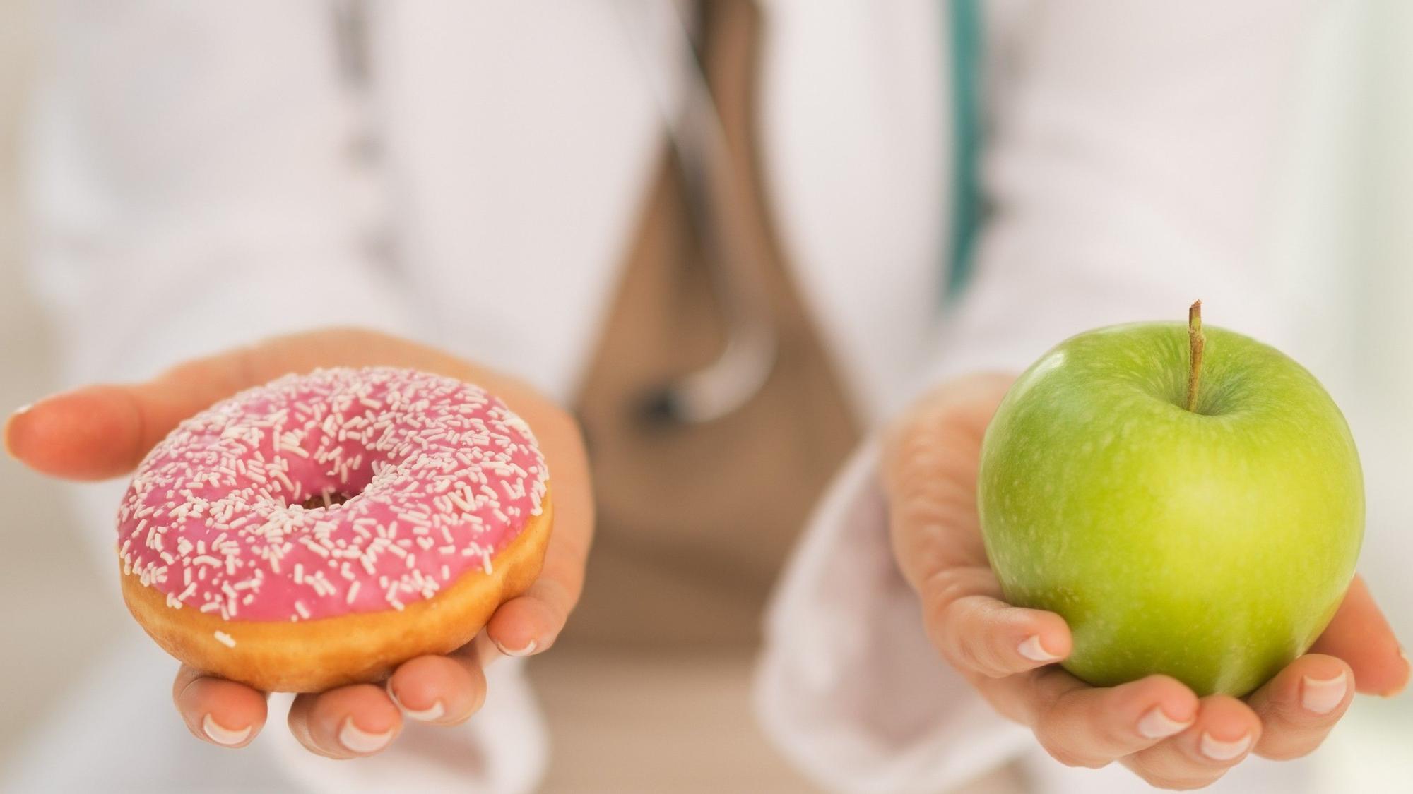 Una doctora sostiene un donut o rosquilla en una mano y una manzana en la otra