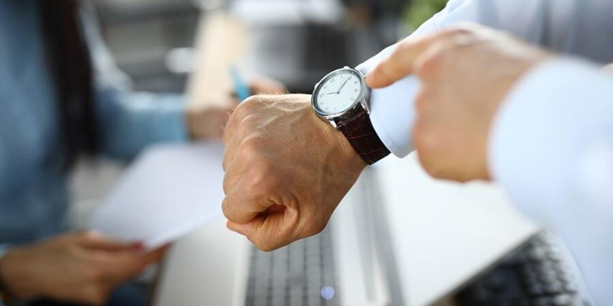 El dedo de un trabajador señalando un reloj a otro, haciendo señal de que ha llegado tarde