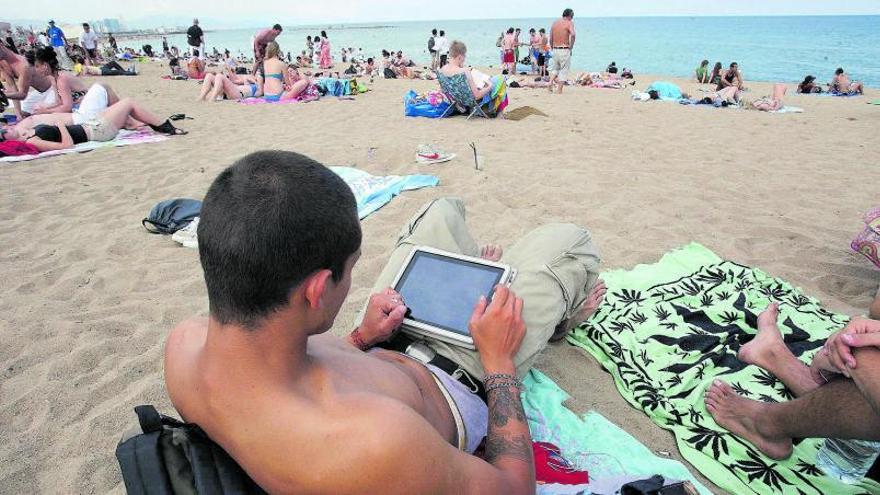 Un joven italiano consulta sus ‘mails’ en una tableta mientras trata de descansar en la playa de la Barceloneta durante sus vacaciones. | ELISENDA PONS
