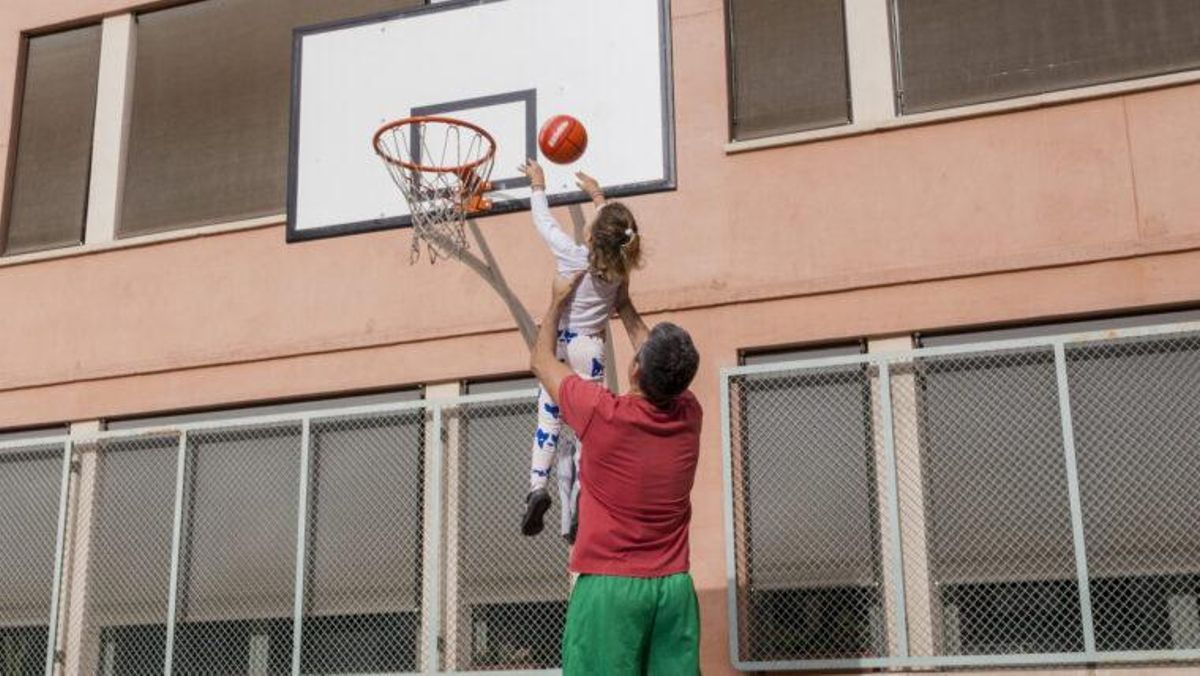 Un padre levanta en brazos a su hija para ayudarla a encestar una pelota de baloncesto.