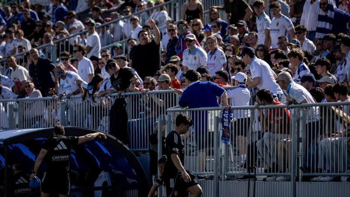 Aficionados zaragocistas de pie hablan con los integrantes del banquillo.