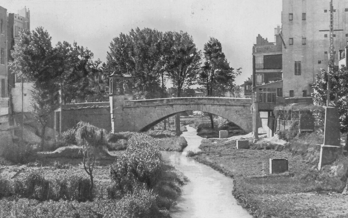 Alzira 1966. El desarrollismo de los años sesenta transformó el perfil de muchas ciudades. Alzira apostó por derribar el histórico Pont de Sant Bernat, cuyos orígenes se remontan al siglo XIII, para levantar una pujante avenida llena de grandes edificios. Del puente solo quedan en pie los casilicios y una parte del pretil reconstruido recientemente.