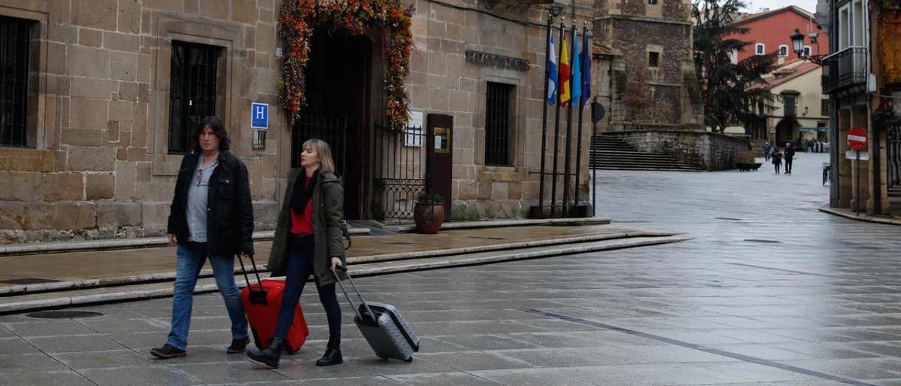 Dos turistas con maletas transitan por El Parche con uno de los hoteles de la ciudad a su espalda.