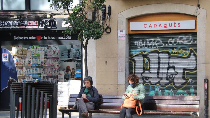 Dues dones assegudes a la plaça de la Vila de Gràcia, davant un restaurant tancat |