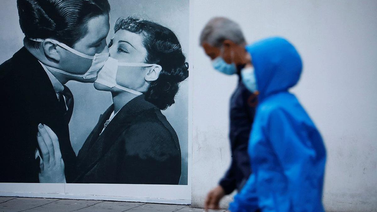 Dos transeúntes pasan junto al cartel de un beso entre dos personas con mascarilla, el domingo 10 de mayo