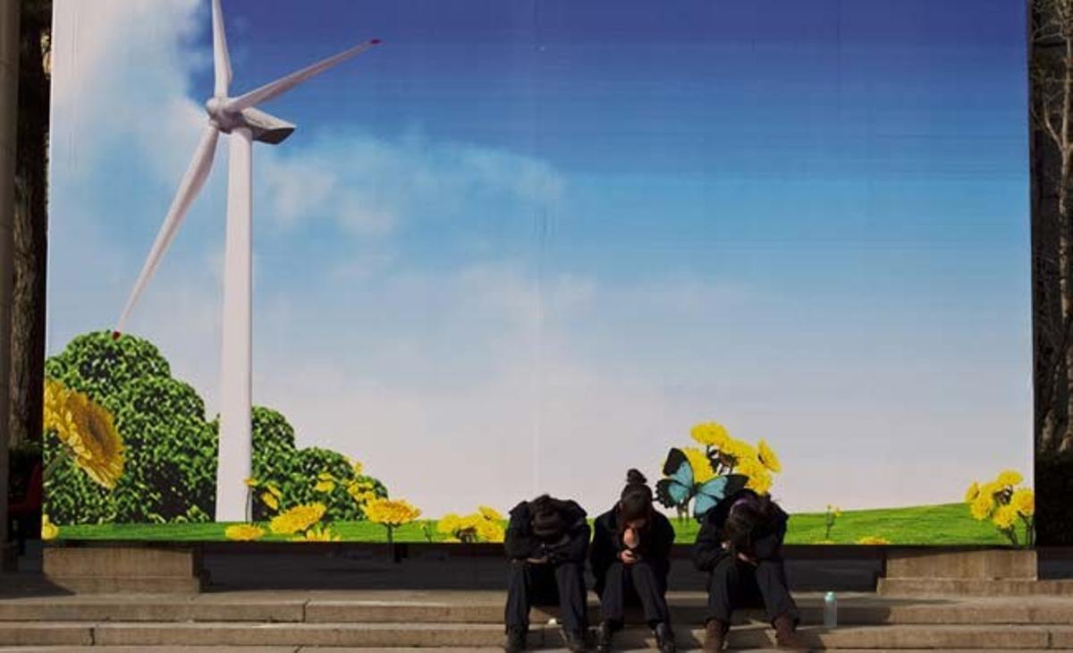 Tres treballadors descansen davant un tauler d’anuncis que mostra un molí de vent, davant la seu d’una exposició de la indústria verda a Pequín (Xina).