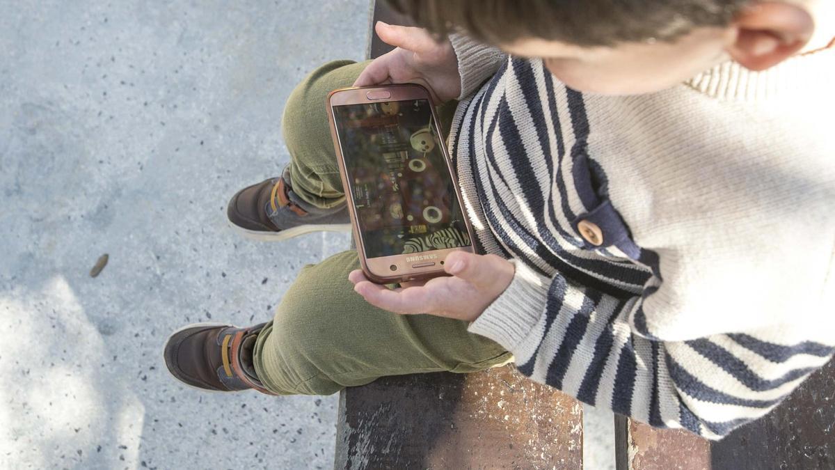 Un niño sentado en un banco del parque con un teléfono móvil en la mano, con el que juega a un videojuego.