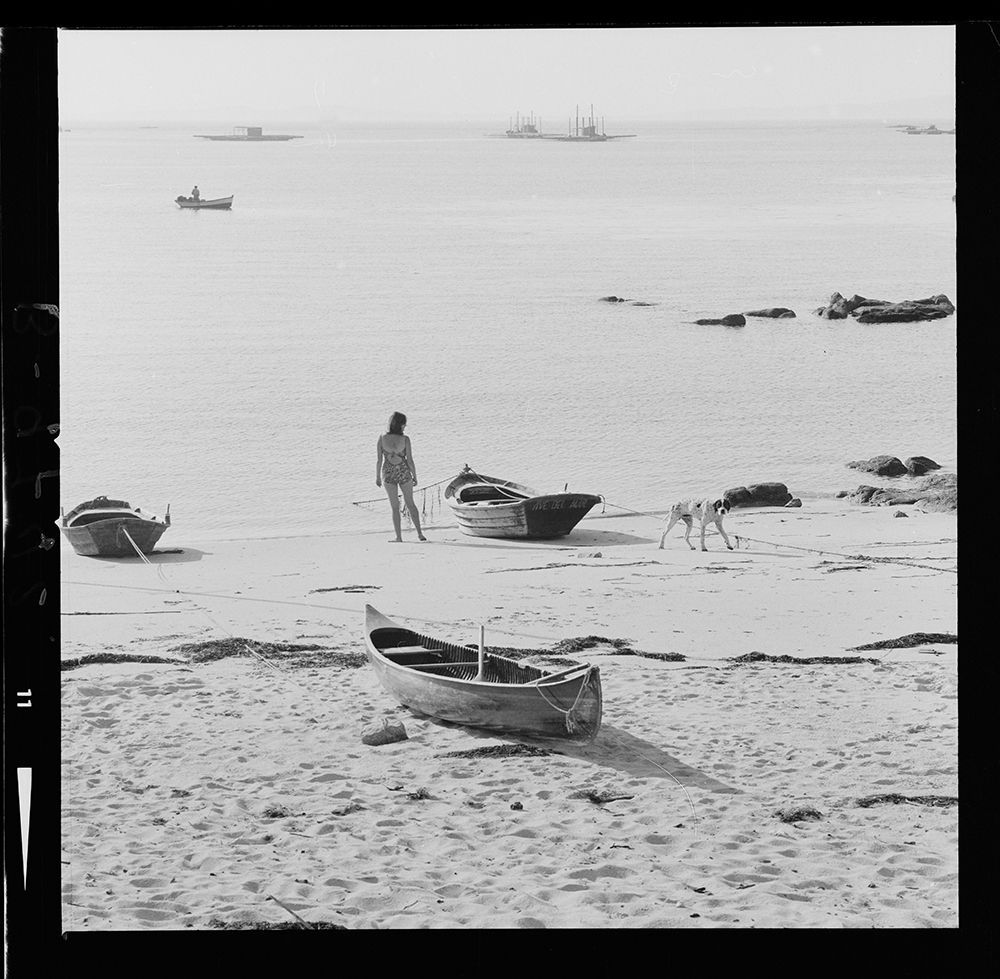 ontevedra. Playas de San Vicente. La Toja [Vista de una playa con barca en la arena y mejilloneras al fondo. En la orilla una mujer en bañador mira hacia el mar y un perro camina por la playa 1967.jpg