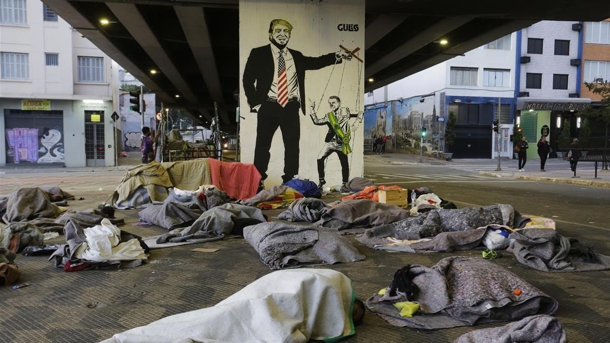 Un grupo de sin techo duerme bajo un puente de Sao Paulo ante una pintada de Bolsonaro como marioneta de Trump.