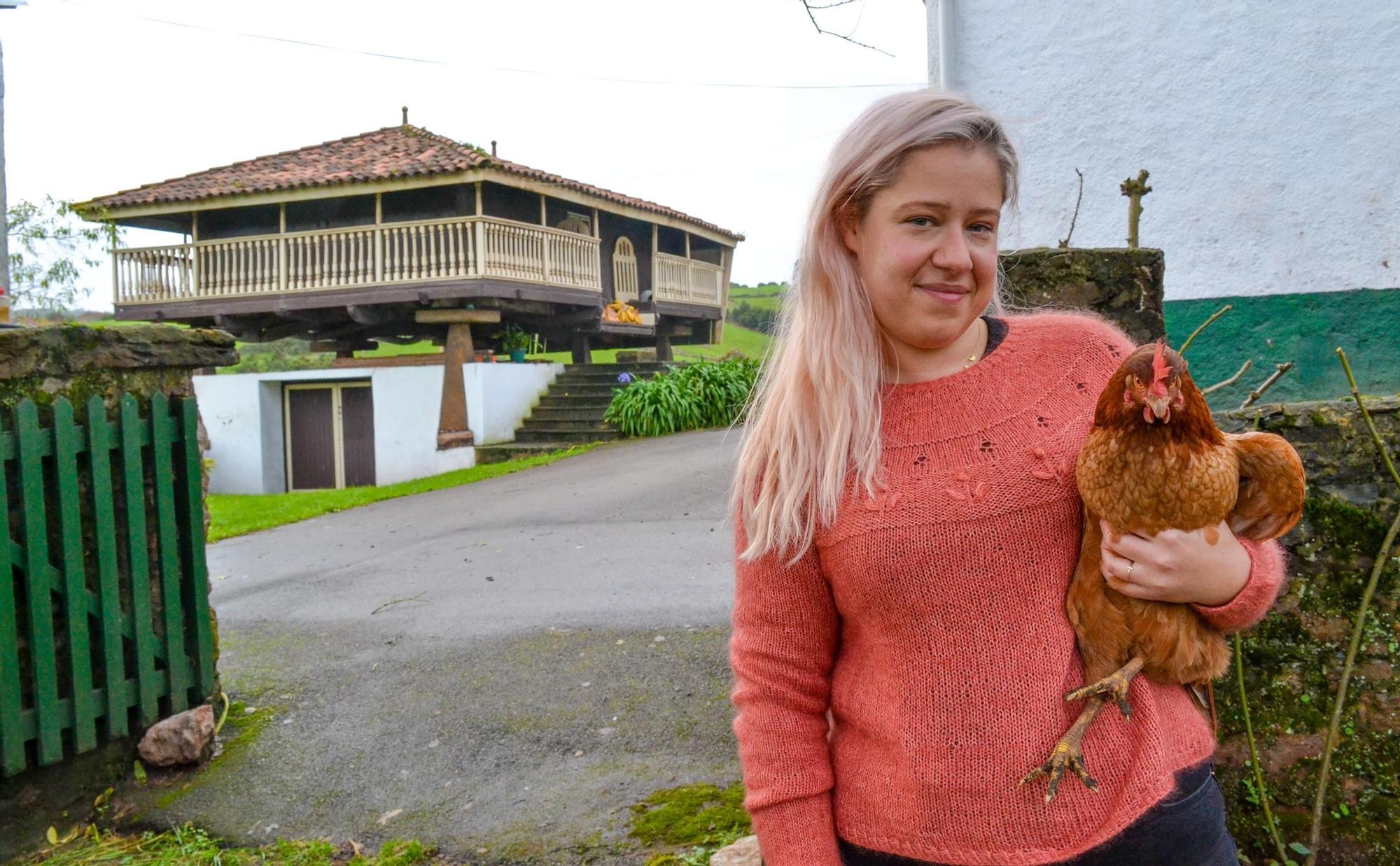 Laura Menéndez posa con una de sus gallinas, vistiendo otro de los jerseis patronados por ella. Al fondo, la panera de la casa.