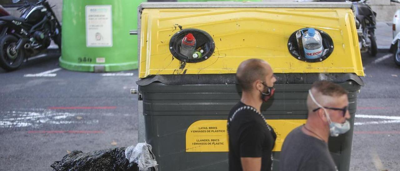 Dos jóvenes pasan junto a un contenedor de recogida de plásticos en Alicante