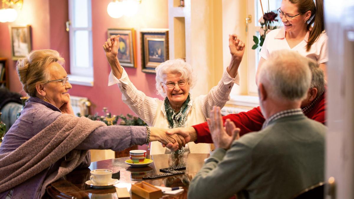 Cuatro amigos mayores disfrutan de un juego de mesa en presencia de una cuidadora.