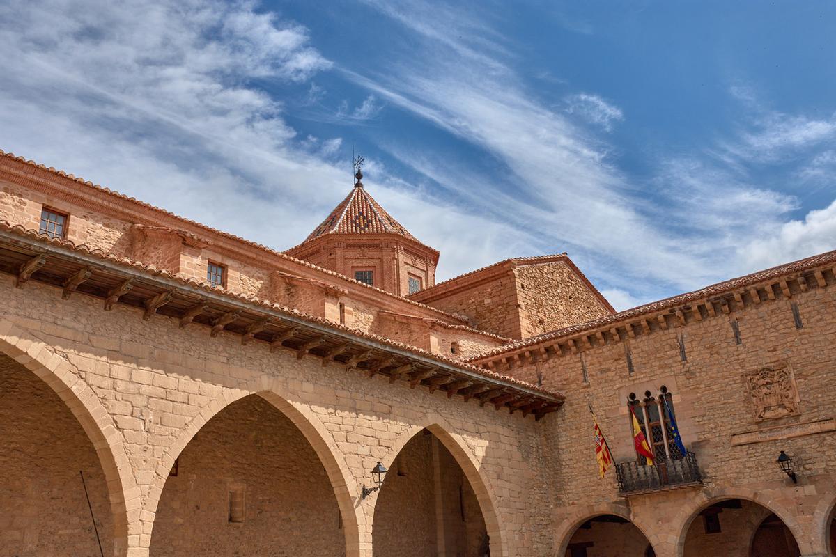 Vista de la plaza de Cristo Rey en Cantavieja con arcos de piedra y una torre histórica bajo un cielo azul en el Alto Maestrazgo