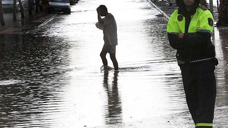 Un vecino cruza una calle de Alicante, completamente inundada, ante la mirada de un Policía Local. A la derecha, vecinos achicando agua en La Albufereta