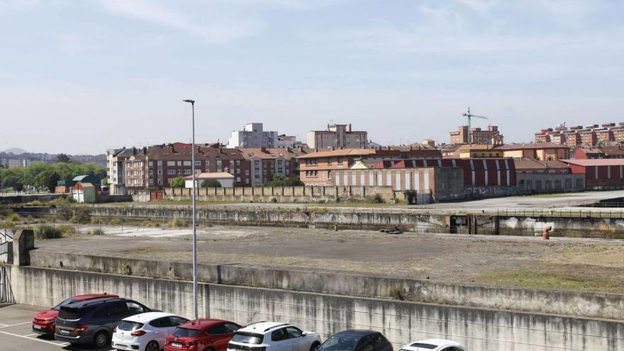 Parte de los terrenos de Naval Gijón, separados por un muro del aparcamiento del Acuario, con edificios del barrio de El Natahoyo al fondo. | ÁNGEL GONZÁLEZ