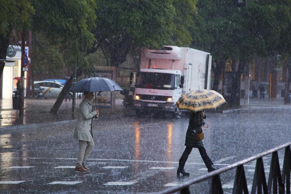 Archivo - Imagen de archivo de dos mujeres cruzando protegiéndose de fuertes lluvias bajo sus respectivos paraguas en un paso de peatones en Sevilla.