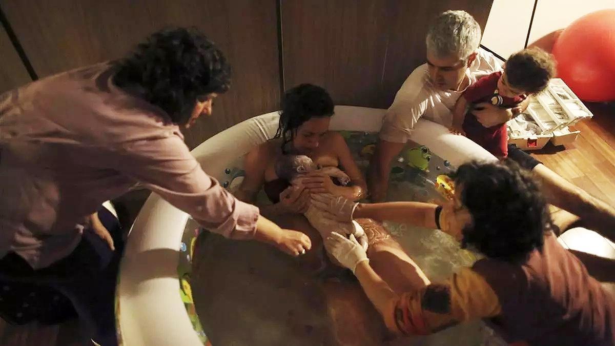 Una mujer da a luz en su casa, rodeada de su familia y otros asistentes, dentro de una piscina de agua caliente.
