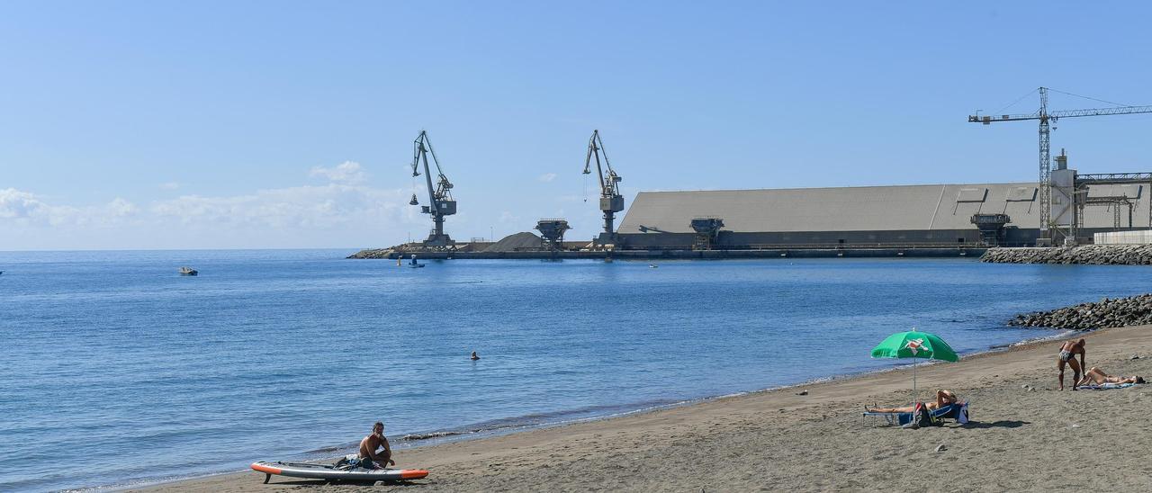 Vecinos y visitantes toman el sol en la bahía de El Pajar con la maquinaria de la cementera en el puerto, al fondo.